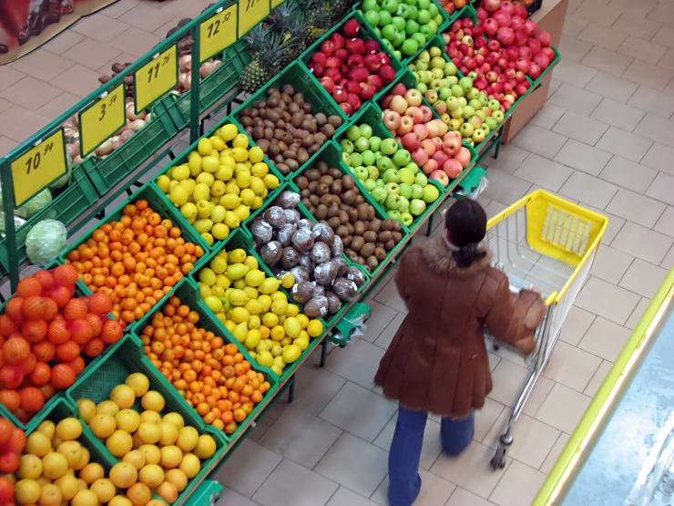 Brightly colored fruits sit in bins inside a grocery store as someone walks by while pushing a grocery cart.